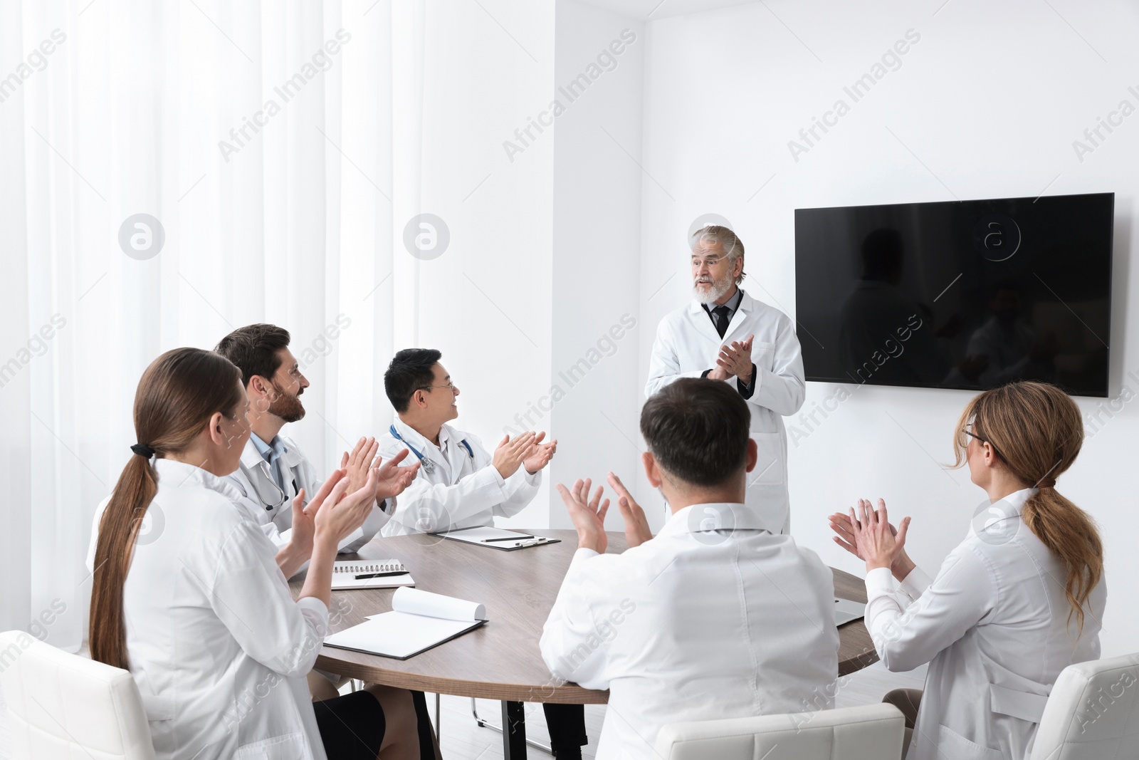 Photo of Team of doctors listening to senior speaker report near tv screen in meeting room. Medical conference