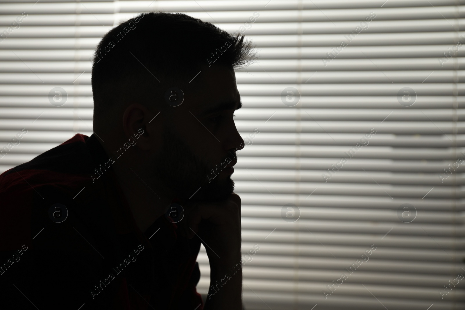 Photo of Silhouette of sad man near closed blinds indoors. Space for text