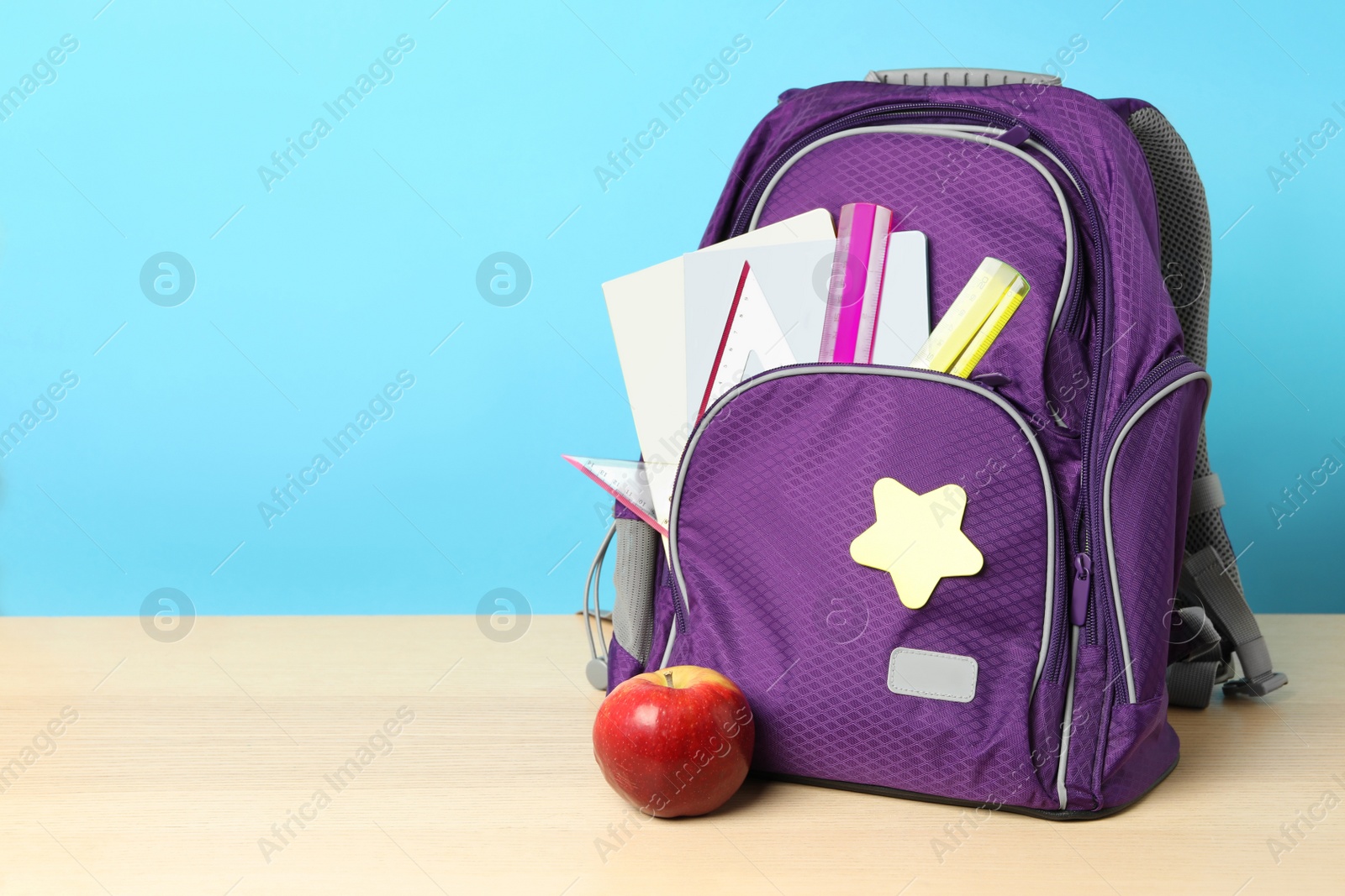 Photo of Stylish backpack with different school stationery and apple on wooden table against light blue background, space for text. Back to school