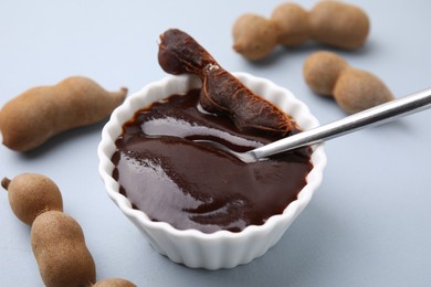 Photo of Bowl of tamarind sauce, spoon and fresh pods on light grey table, closeup