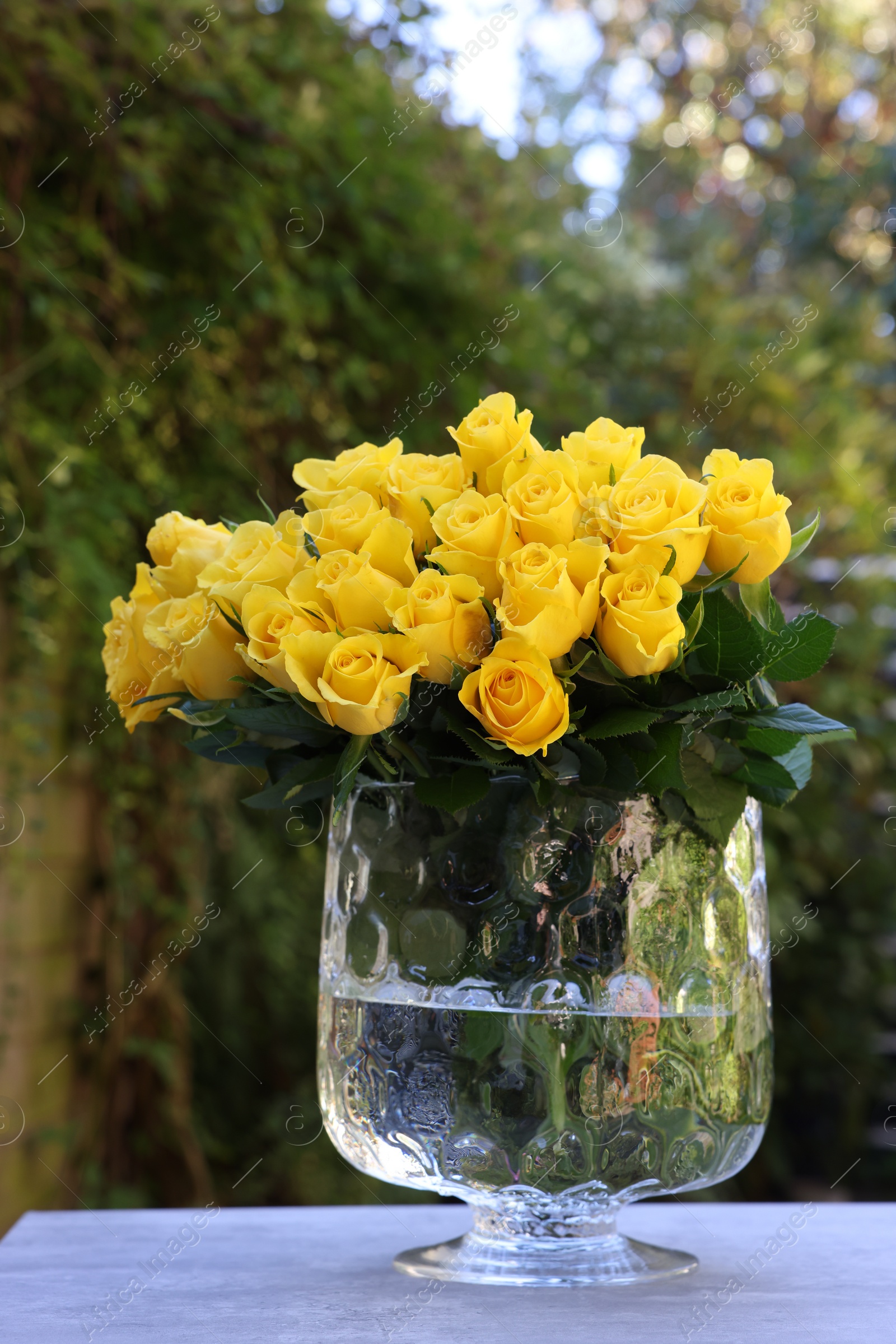 Photo of Beautiful bouquet of yellow roses in glass vase on light table outdoors