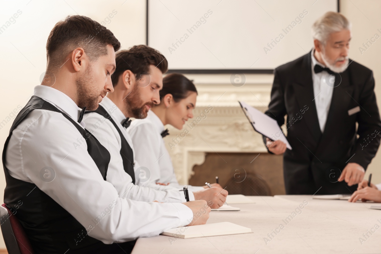 Photo of Senior man in formal suit teaching trainees indoors. Professional butler courses