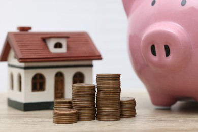 Photo of House model, piggy bank and stacked coins on light wooden table, closeup