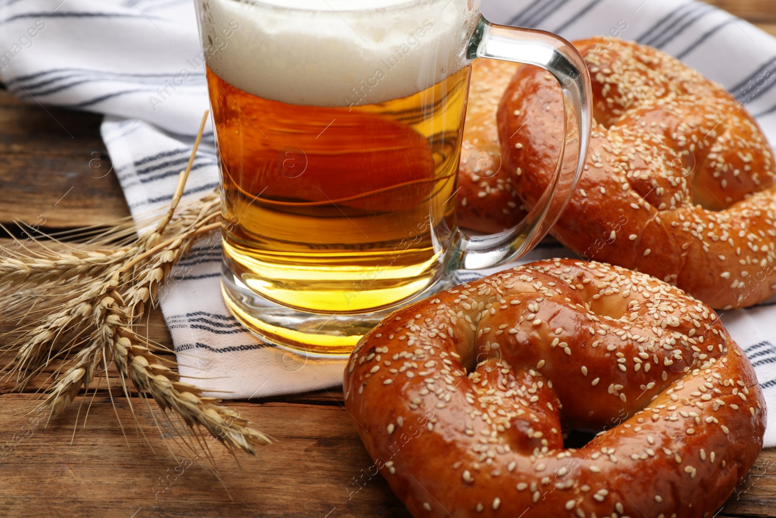 Photo of Tasty pretzels, glass of beer and wheat spikes on wooden table, closeup