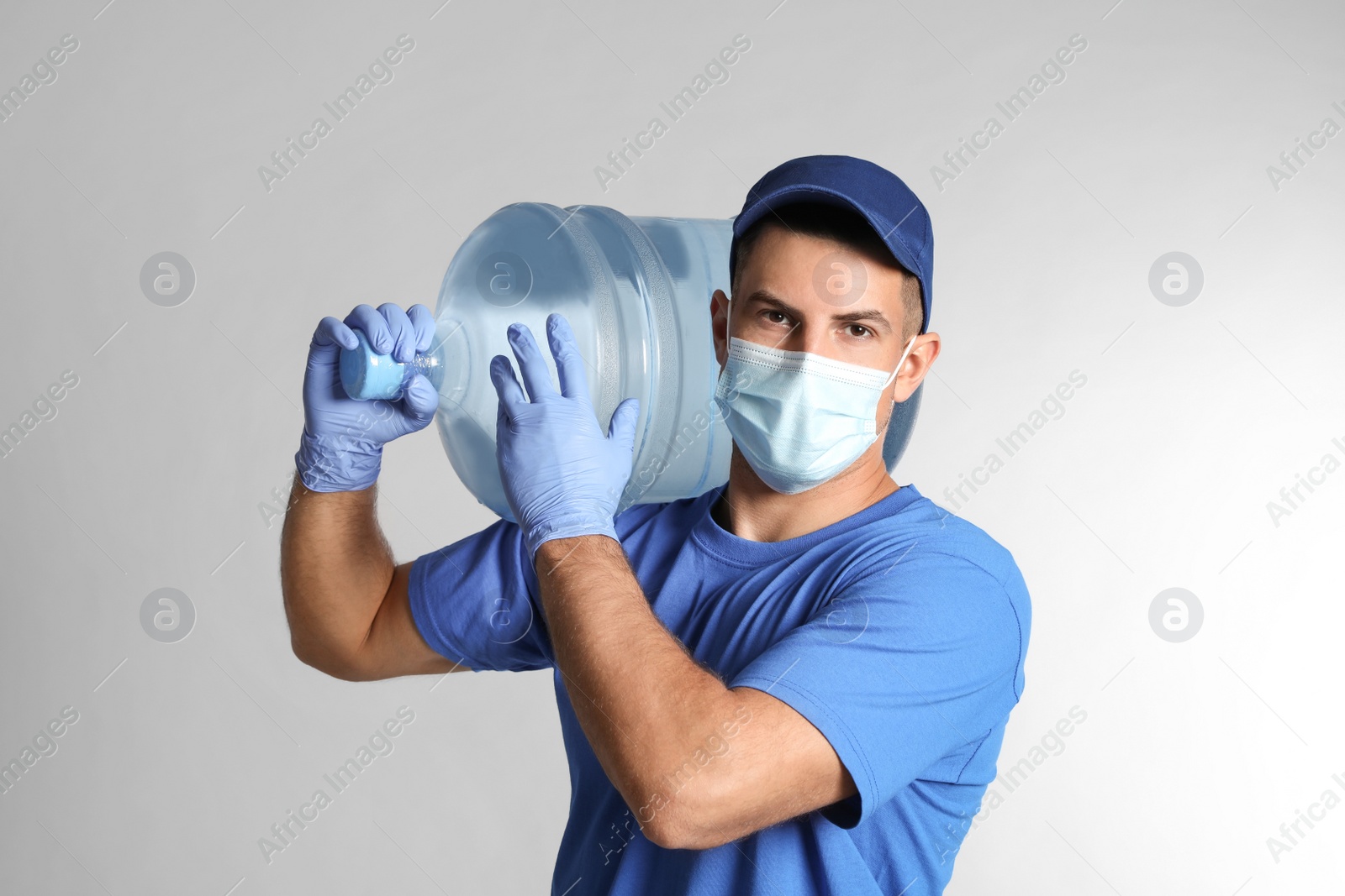 Photo of Courier in face mask with bottle of cooler water on grey background. Delivery during coronavirus quarantine