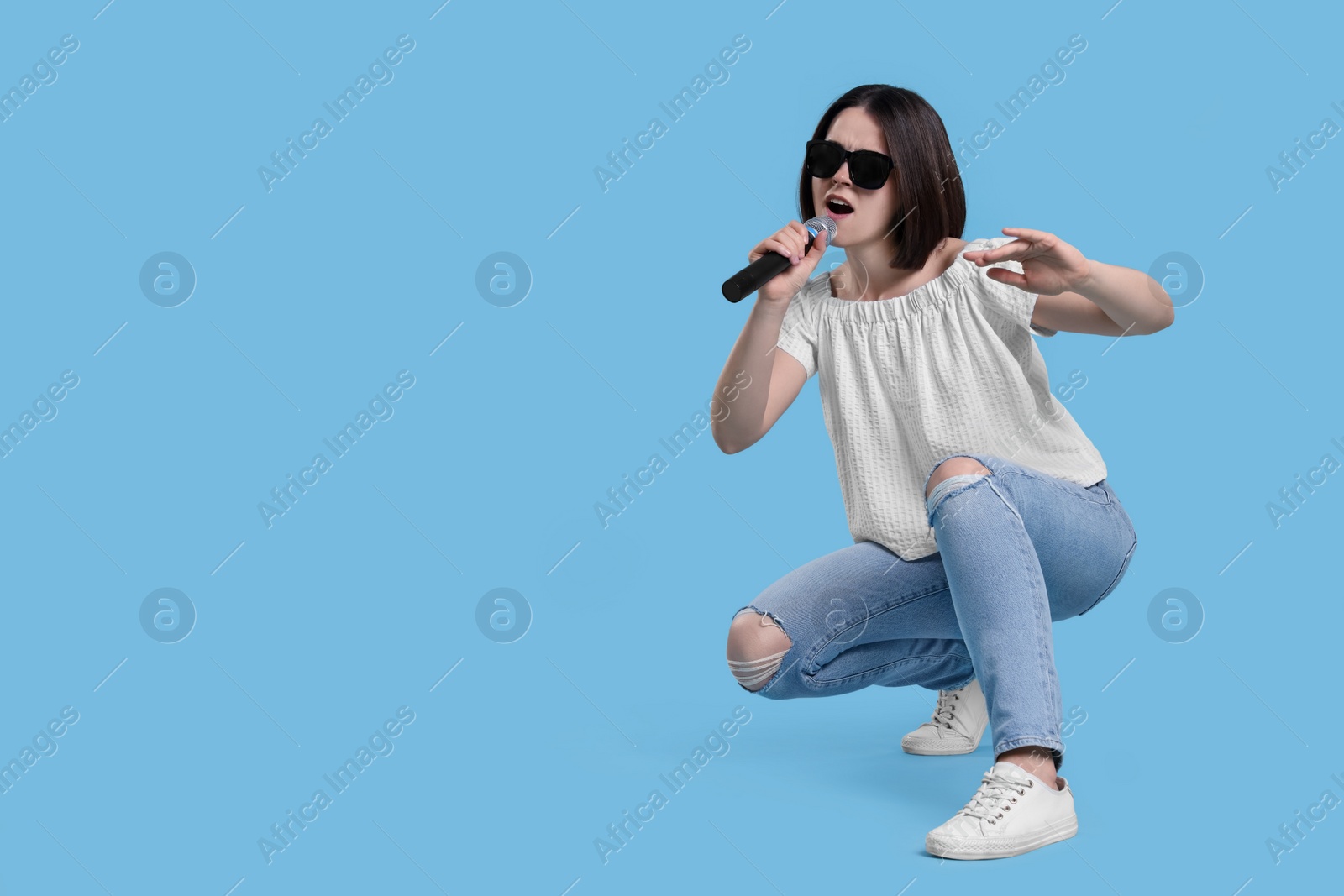 Photo of Beautiful young woman with sunglasses and microphone singing on light blue background, space for text