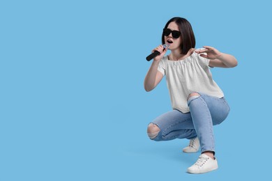 Beautiful young woman with sunglasses and microphone singing on light blue background, space for text