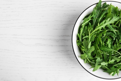 Fresh arugula in bowl on white wooden table, top view. Space for text