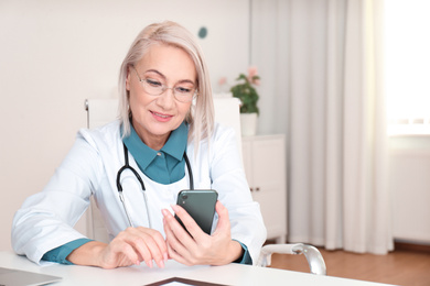 Photo of Mature female doctor with smartphone at table in office