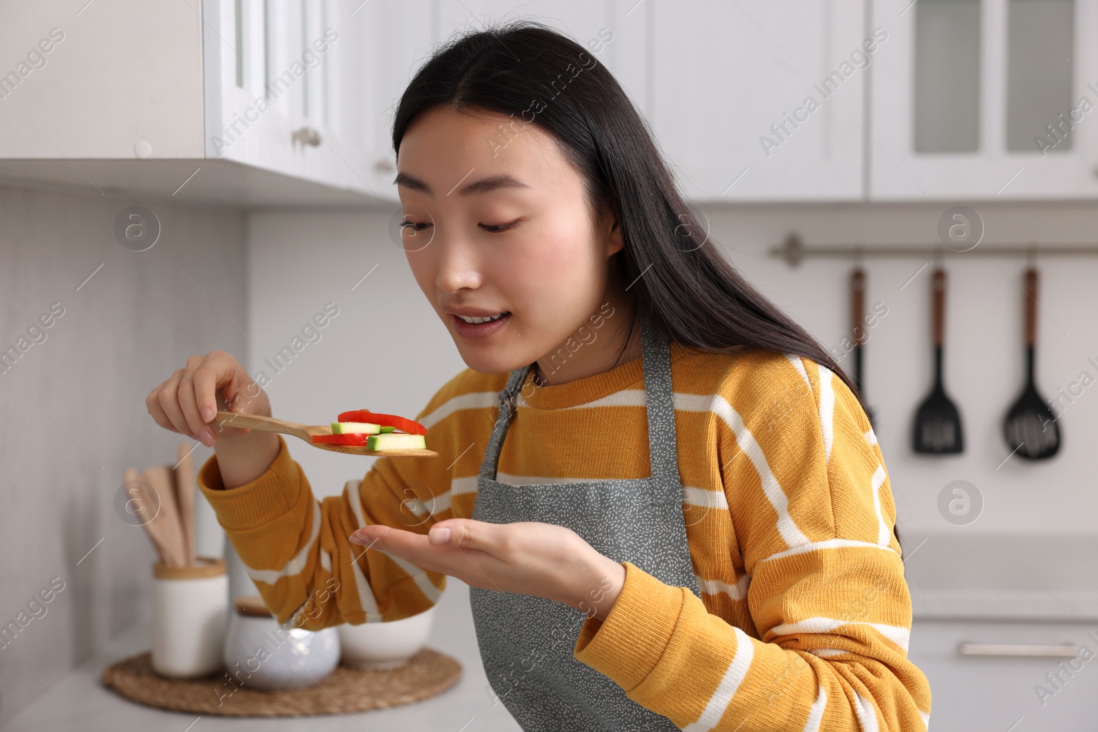 Photo of Beautiful woman cooking and tasting vegetable dish in kitchen