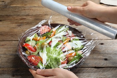 Woman putting plastic food wrap over bowl of fresh salad at wooden table, closeup