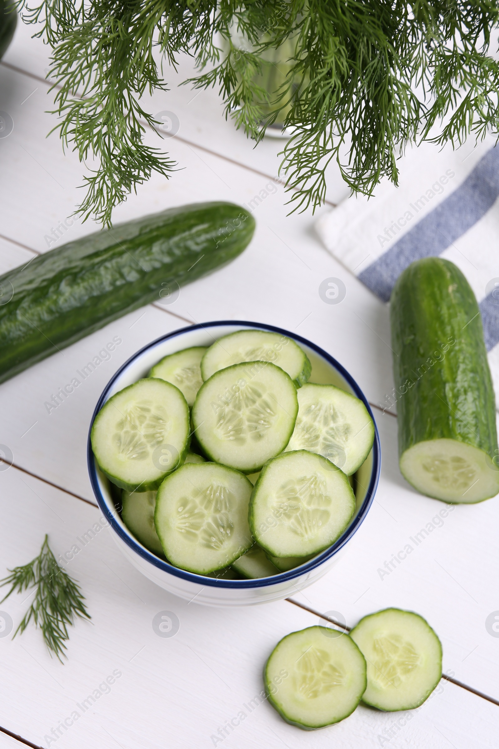 Photo of Cut cucumber in bowl, fresh vegetables and dill on white wooden table, above view