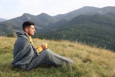 Man with cup of drink in sleeping bag enjoying mountain landscape, space for text