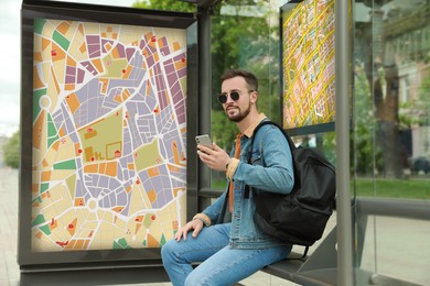 Image of Young man with smartphone and backpack waiting for public transport at bus stop
