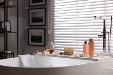 Wooden tray with wine, toiletries and flower petals on bathtub in bathroom