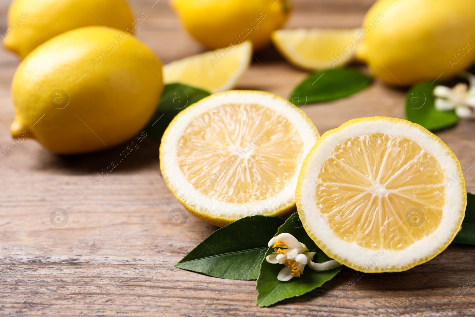 Photo of Many fresh ripe lemons with green leaves and flowers on wooden table, closeup