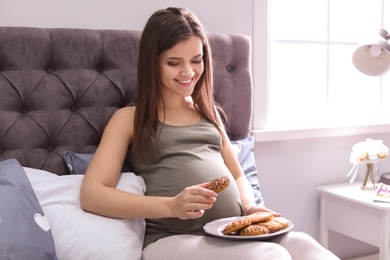 Photo of Young pregnant woman holding plate with cookies on bed