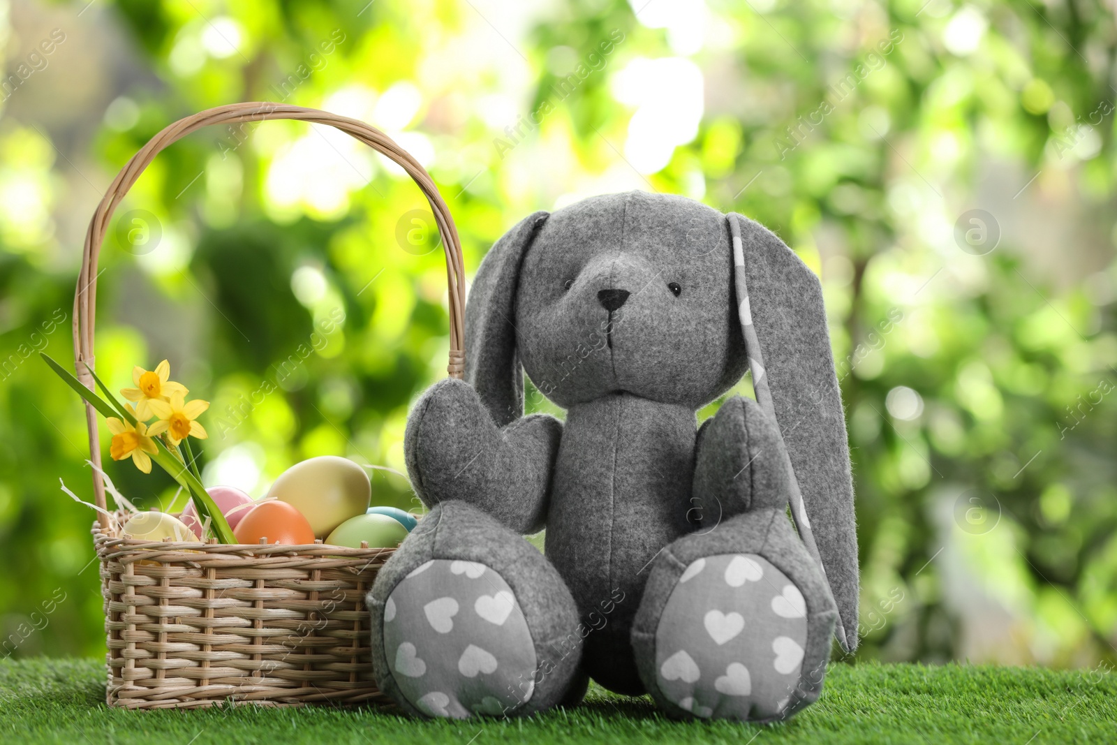 Photo of Cute Easter bunny toy and wicker basket with dyed eggs on green grass against blurred background