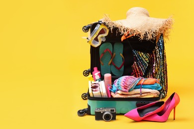 Open suitcase with clothes, beach accessories and shoes on yellow background, space for text. Summer vacation
