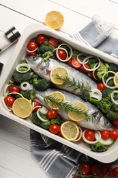 Photo of Raw fish with vegetables in baking dish and lemon on white wooden table, flat lay