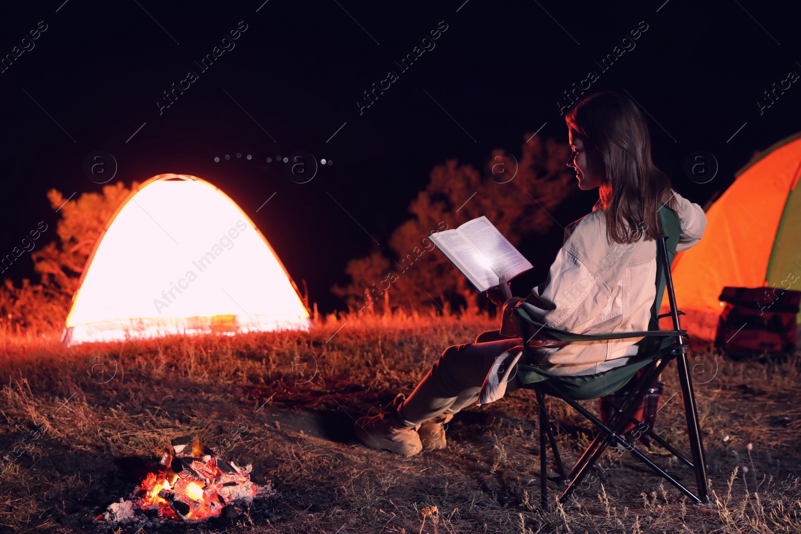 Photo of Young woman with flashlight reading book near bonfire at night. Camping season