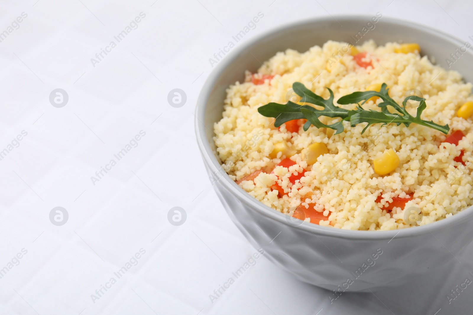 Photo of Tasty couscous with pepper, corn and arugula in bowl on white tiled table, closeup. Space for text