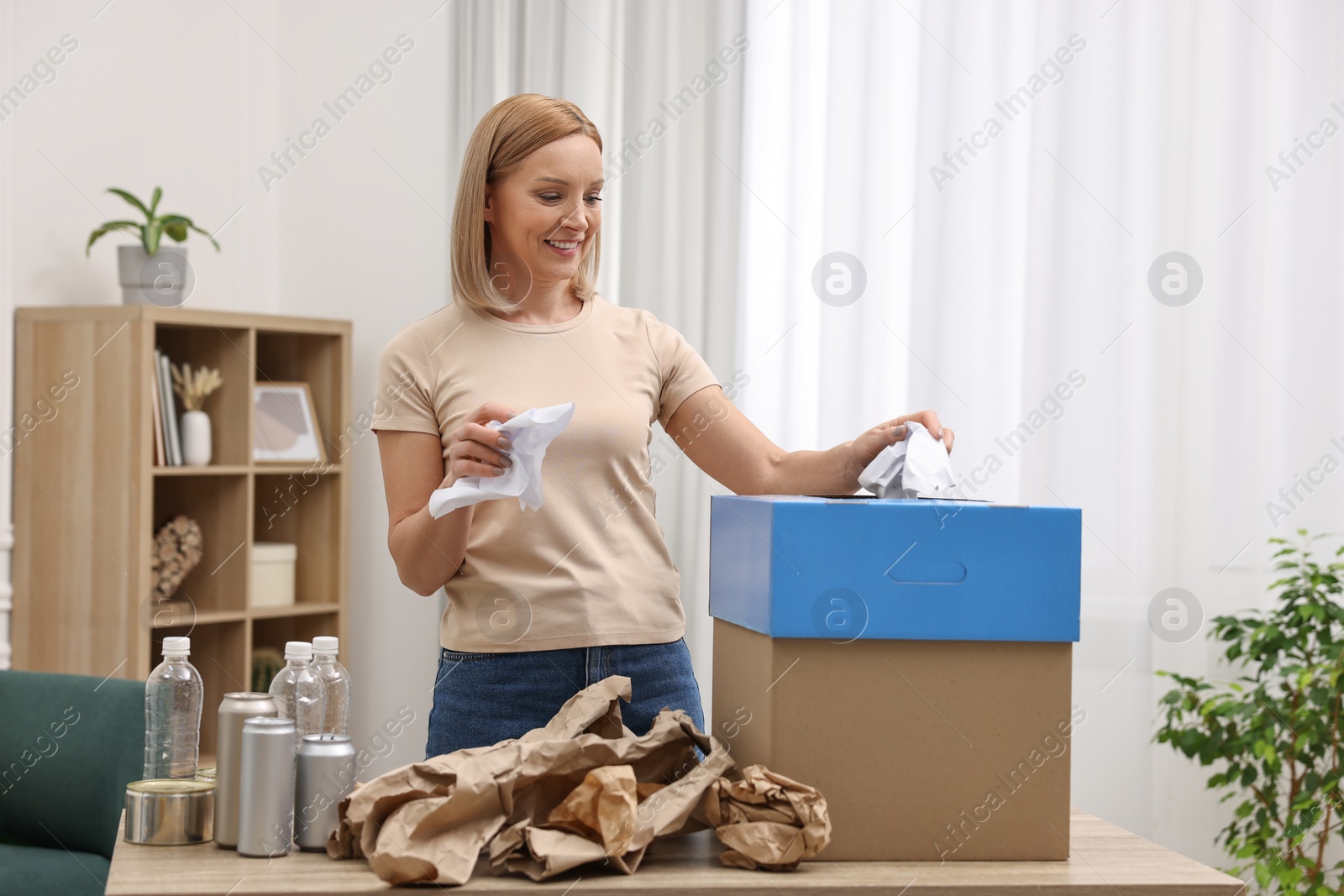 Photo of Garbage sorting. Smiling woman throwing crumpled paper into cardboard box in room