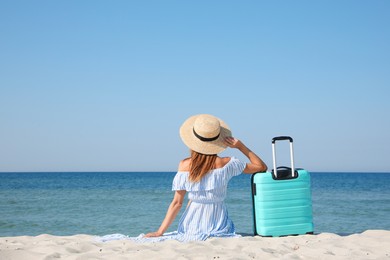Photo of Woman with suitcase sitting on sandy beach near sea, back view