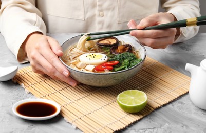 Photo of Woman eating delicious ramen with chopsticks at grey table, closeup. Noodle soup