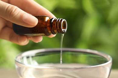 Woman pouring essential oil from glass bottle into bowl against blurred green background, closeup