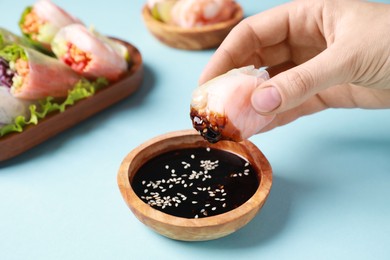 Photo of Woman dipping delicious spring roll wrapped in rice paper into soy sauce on light blue background, closeup