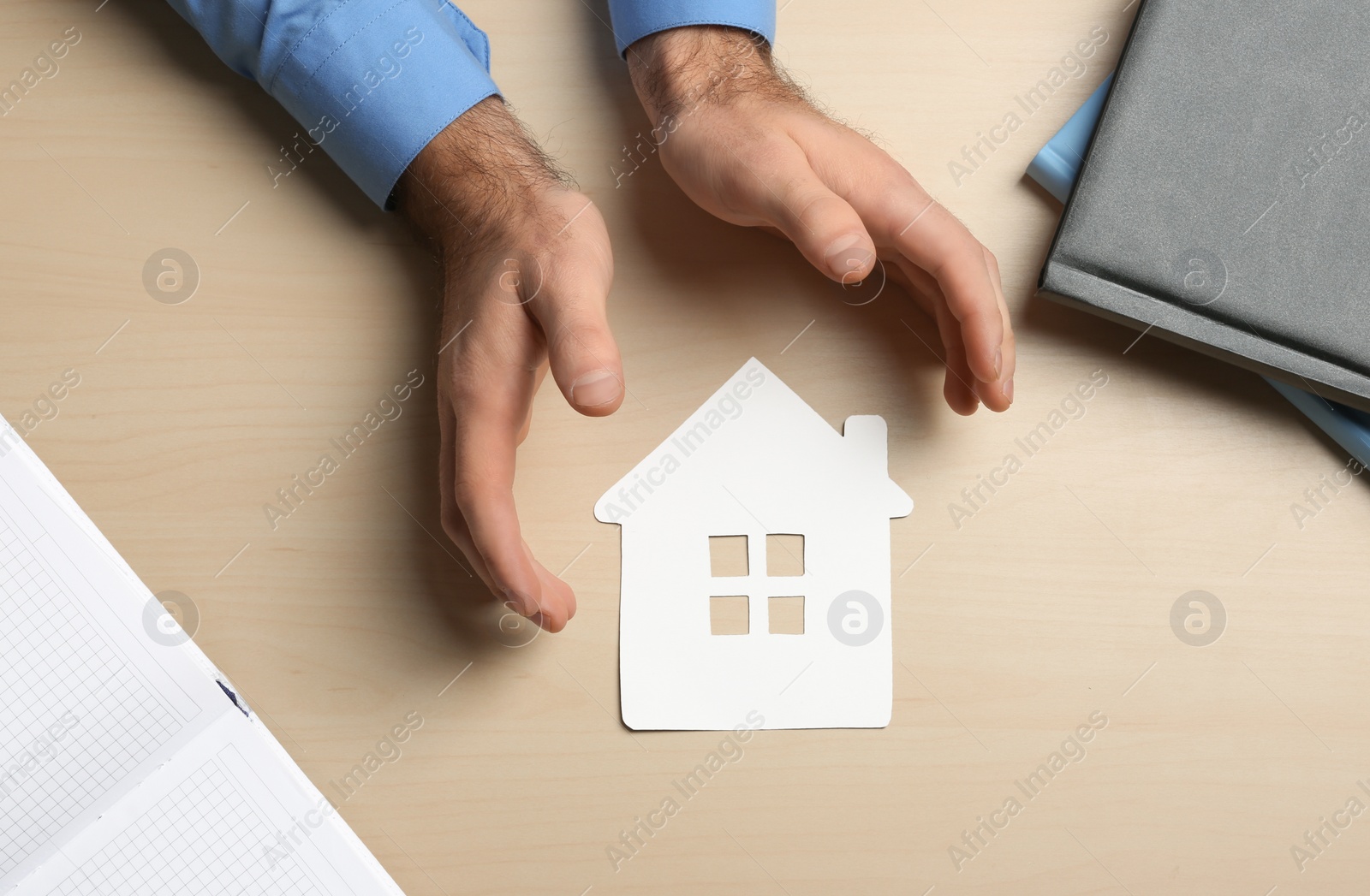 Photo of Man holding hands near paper silhouette of house on wooden background, top view. Home insurance