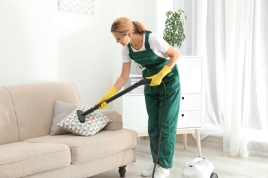 Photo of Female janitor removing dirt from sofa cushion with steam cleaner in room