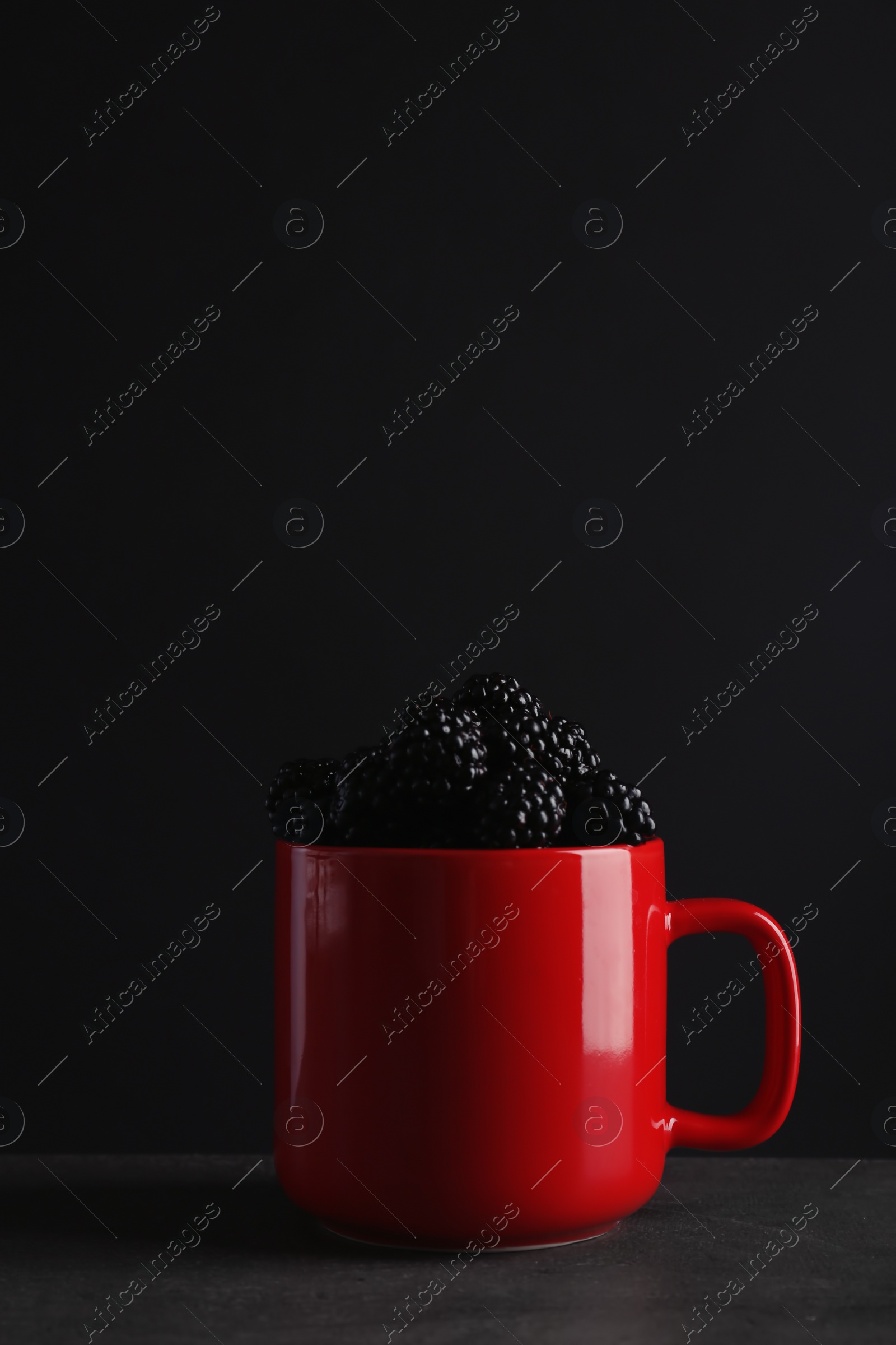 Photo of Mug with fresh blackberry on gray table against dark background. Space for text