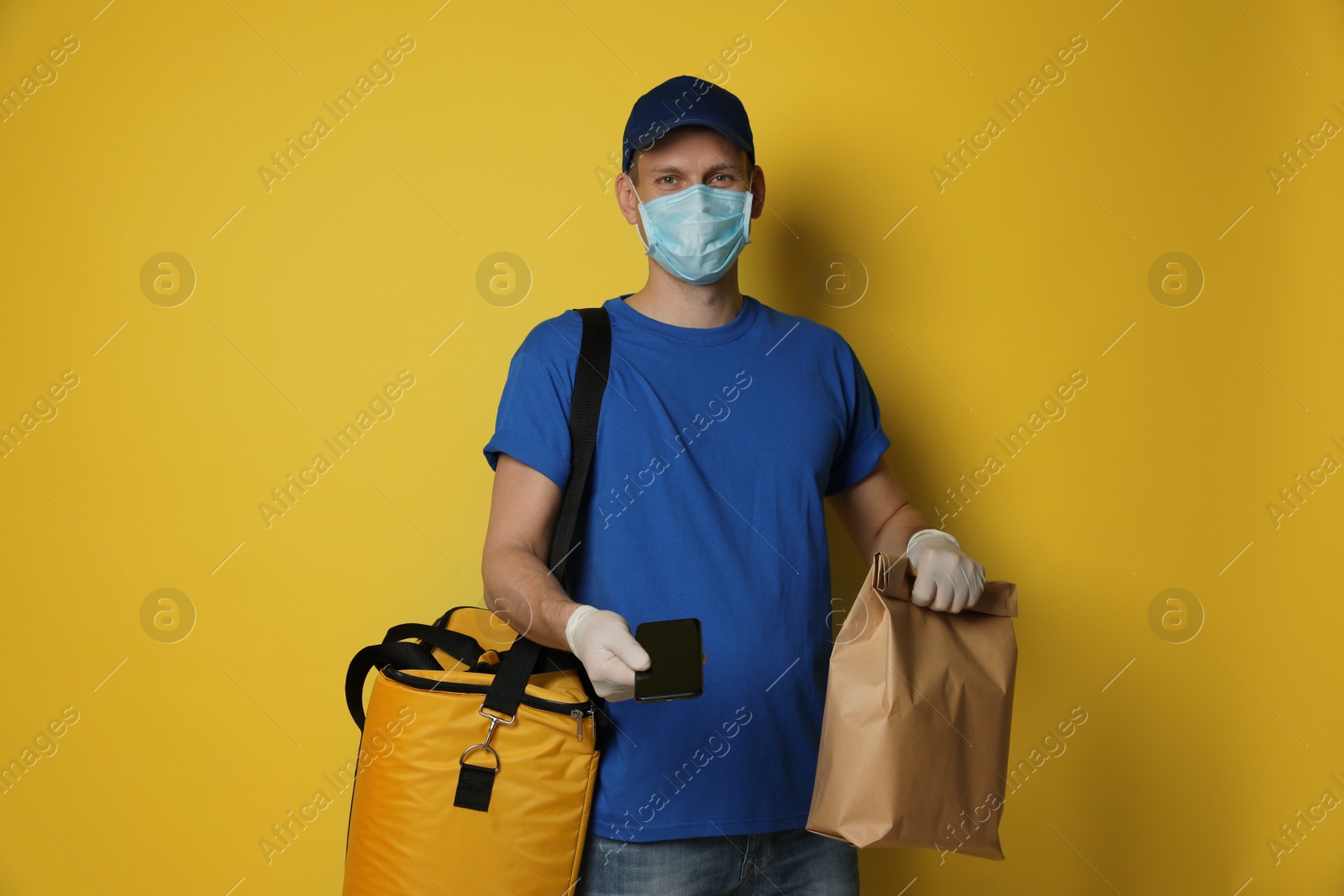 Photo of Courier in protective mask holding order and smartphone for contactless payment on yellow background. Food delivery service during coronavirus quarantine
