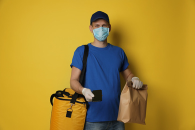 Courier in protective mask holding order and smartphone for contactless payment on yellow background. Food delivery service during coronavirus quarantine