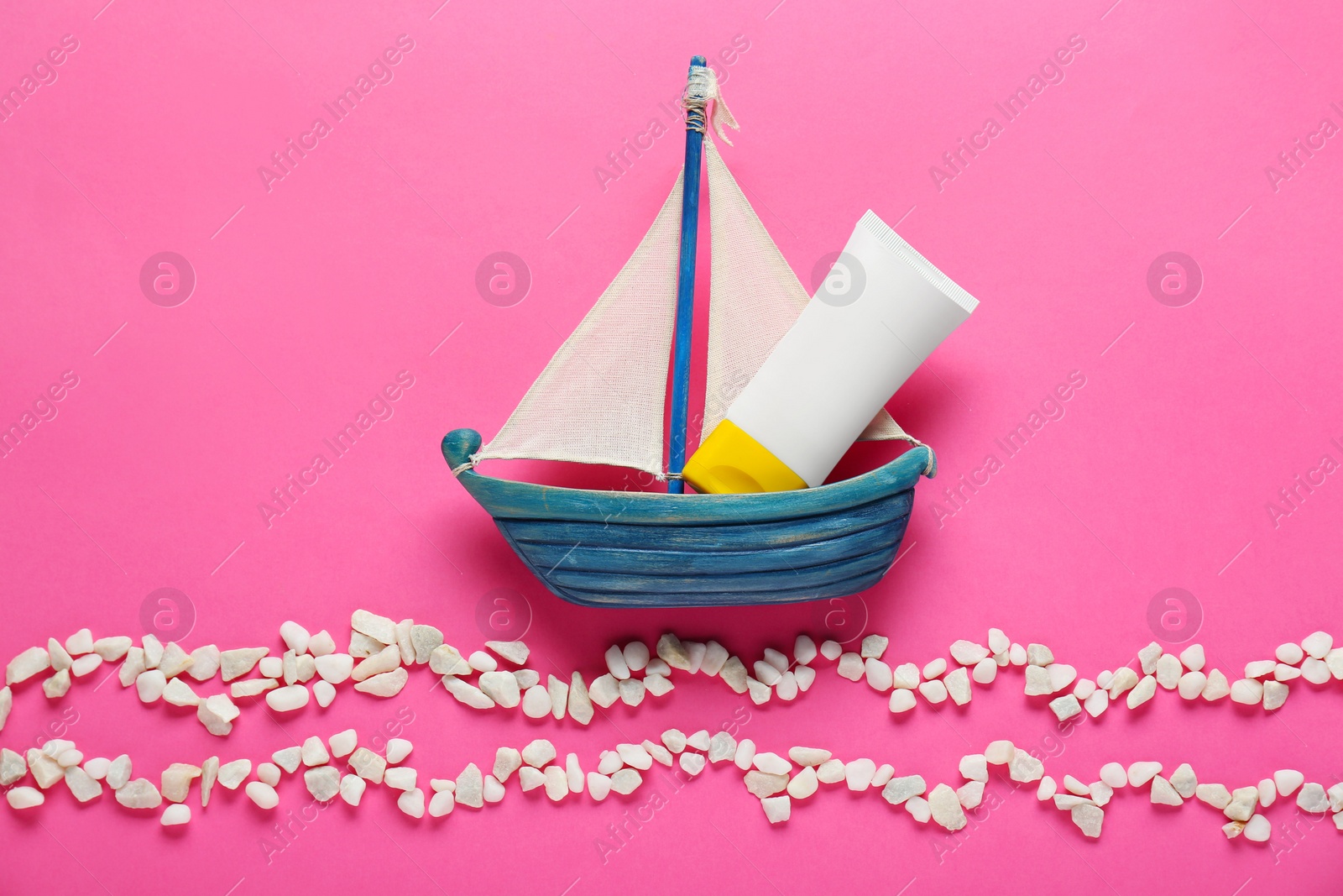 Photo of Suntan cream in toy sailboat and white marble pebbles on pink background, flat lay