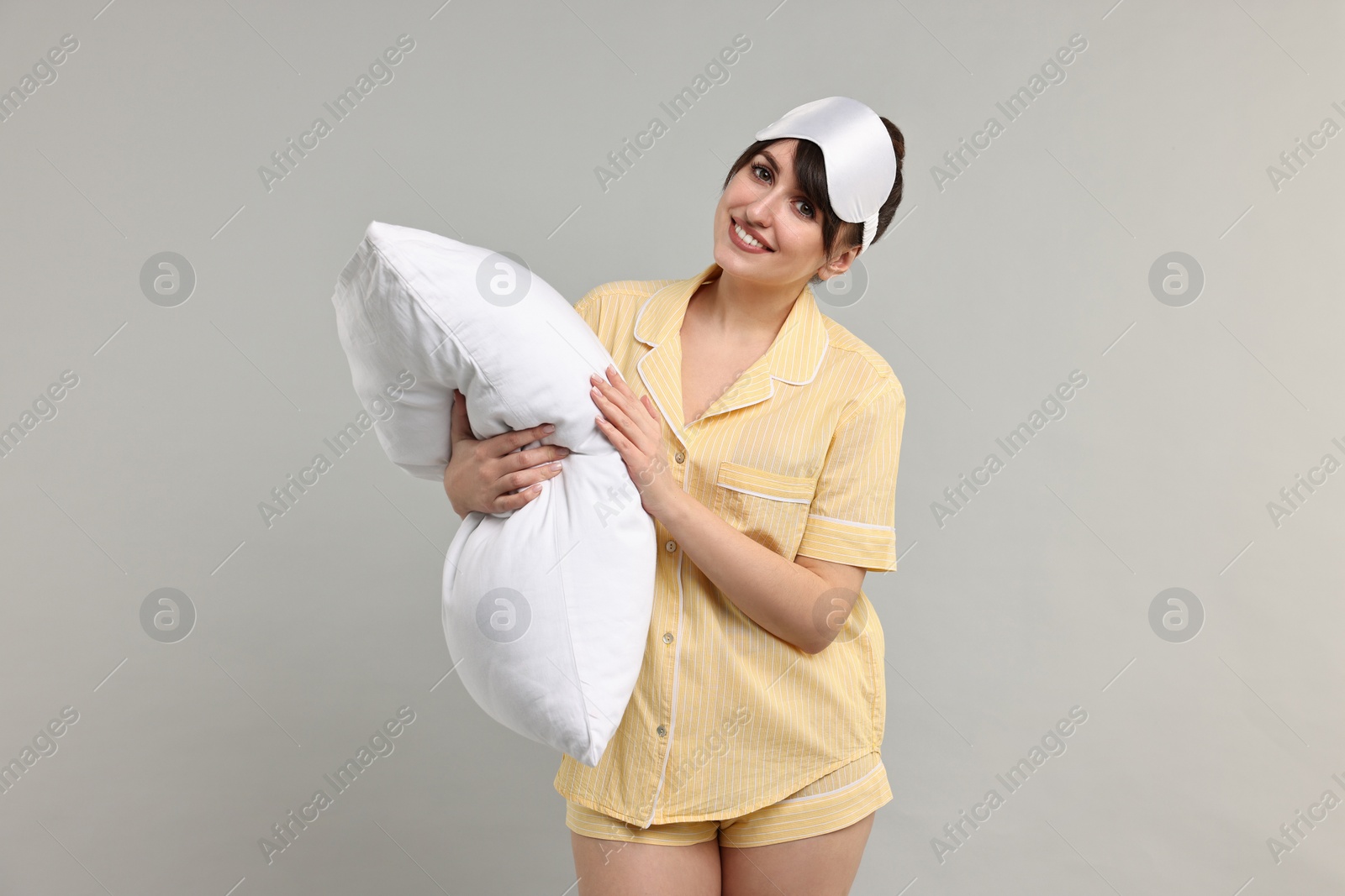 Photo of Happy woman in pyjama and sleep mask holding pillow on grey background