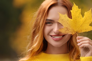 Photo of Smiling woman covering eye with autumn leaf outdoors, closeup. Space for text