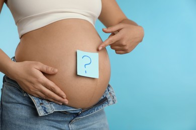 Pregnant woman with sticky note on belly against light blue background, closeup. Choosing baby name