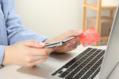 Online payment. Woman using credit card and smartphone near laptop at white wooden table indoors, closeup