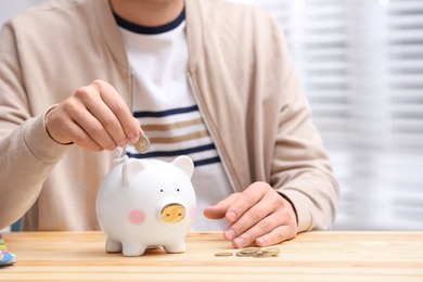 Photo of Man putting coin into piggy bank at wooden table, closeup