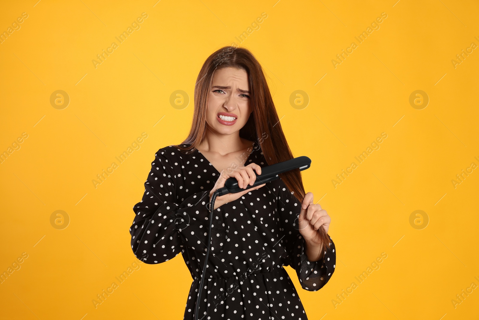 Photo of Stressed young woman with flattening iron on yellow background. Hair damage