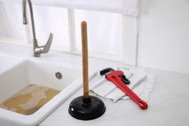 Photo of Plunger, pipe wrench and towel on kitchen counter near clogged sink