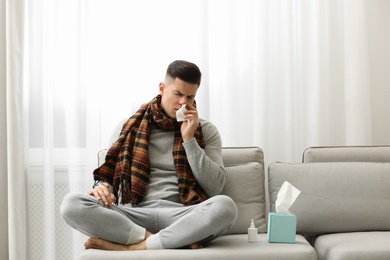 Photo of Ill man sitting near nasal spray and box of paper tissues on sofa at home