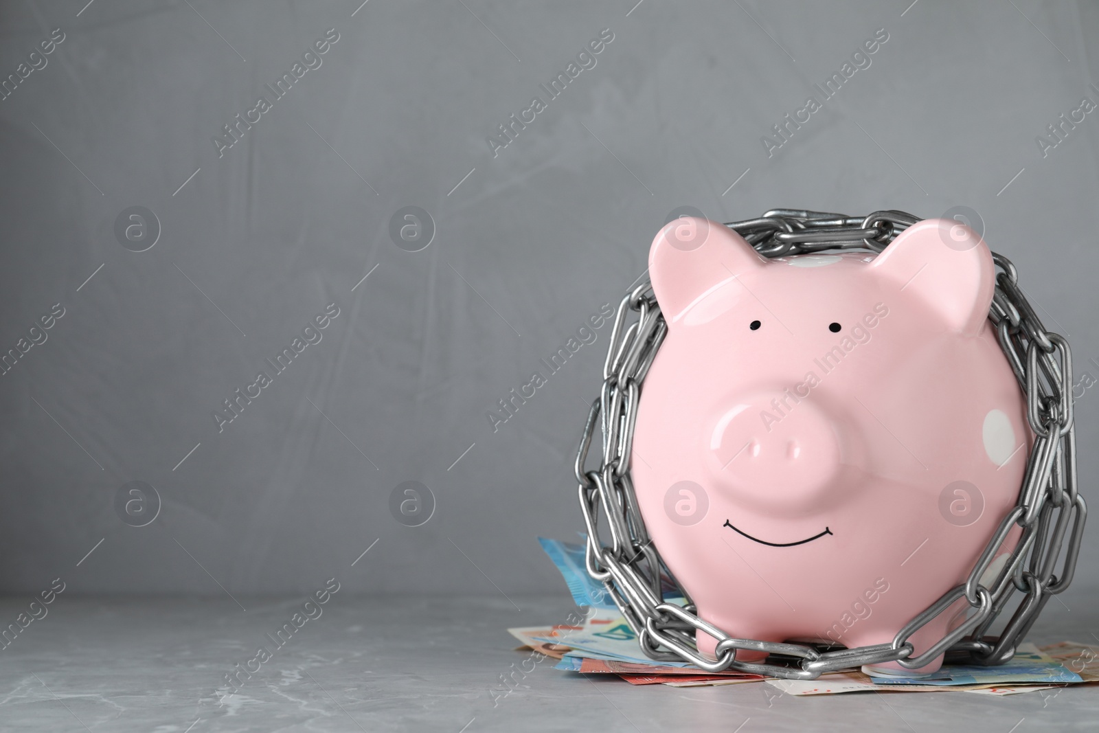 Photo of Piggy bank with steel chain and banknotes on grey marble table, space for text. Money safety concept