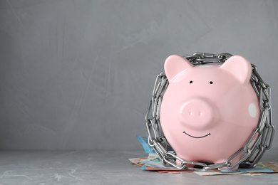 Photo of Piggy bank with steel chain and banknotes on grey marble table, space for text. Money safety concept