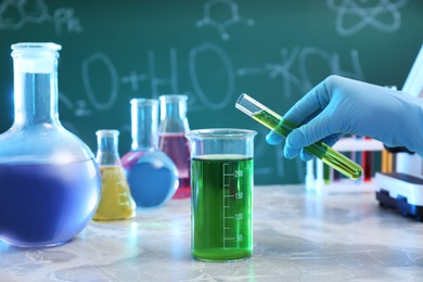 Photo of Scientist pouring liquid into beaker at table against chalkboard, closeup. Chemistry glassware