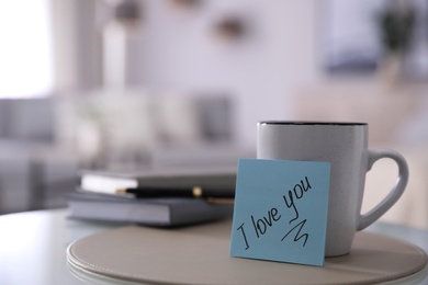 Photo of Paper note with handwritten words I Love You and cup on table indoors. Space for text