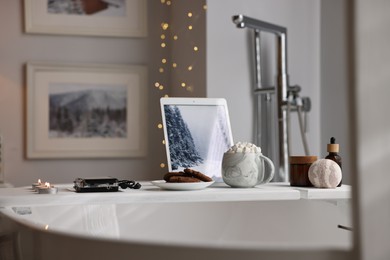 White wooden tray with tablet, spa products and burning candles on bathtub in bathroom